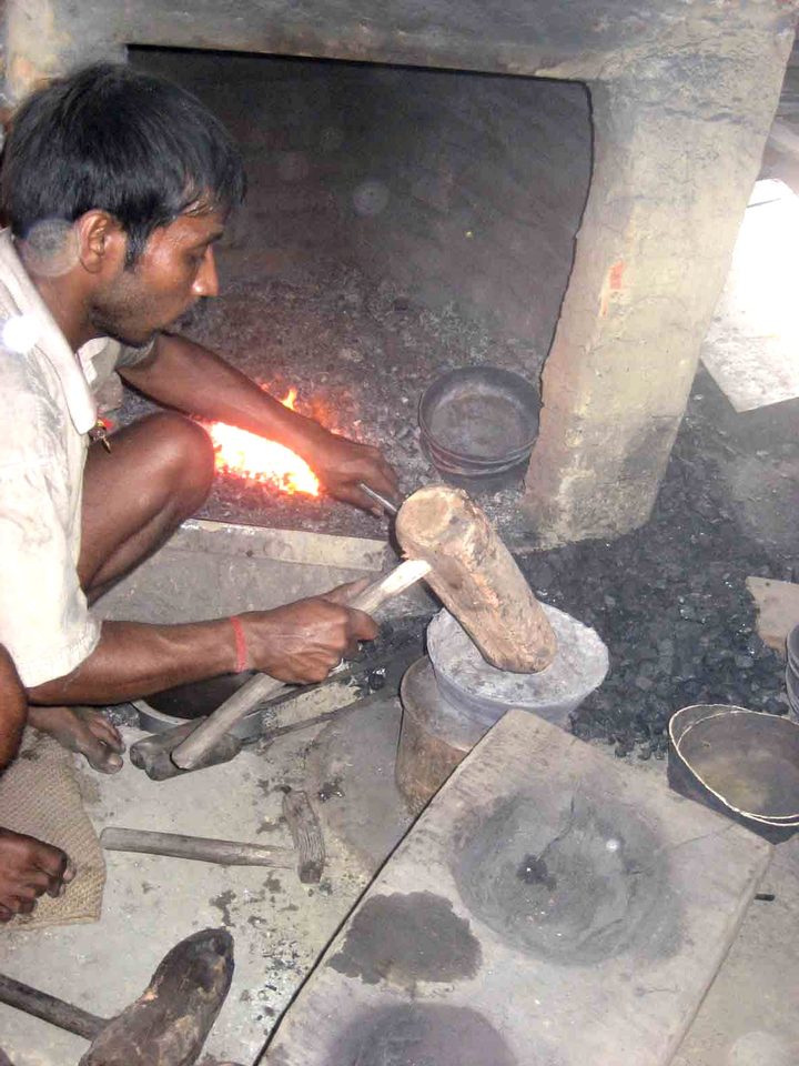 Schmied hält ein glühendes Metallstück mit der Zange über die Esse